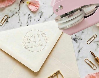 May include: Cream envelope with a circular embossed design showing the initials "KJ" and the date "21.07.2026". A pink paper punch, gold paper clips, and a gold pen are also visible. Pink flowers and petals are scattered around.