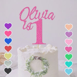 May include: Pink glitter cake topper with the name Olivia and the number 1. The topper is on a white cake with a green wreath.