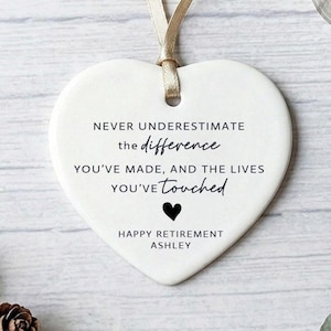 May include: A white heart-shaped ceramic ornament with a beige ribbon. The ornament features black text: "NEVER UNDERESTIMATE the difference YOU'VE MADE, AND THE LIVES YOU'VE touched." A black heart and "HAPPY RETIREMENT ASHLEY" are also present.