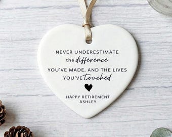 May include: A white heart-shaped ceramic ornament with a beige ribbon. The ornament features black text: "NEVER UNDERESTIMATE the difference YOU'VE MADE, AND THE LIVES YOU'VE touched." A black heart and "HAPPY RETIREMENT ASHLEY" are also present.