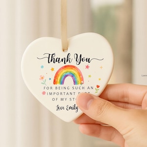 May include: A cream-coloured heart-shaped ceramic ornament with a rainbow design and the words "Thank You." It has a gold ribbon for hanging. Text includes "For being such an important... Love Emily."