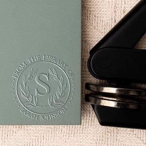 May include: A grey self-inking stamp with the text "From the Library of Sarah Johnson" and a decorative wreath with the letter "S" in the centre.
