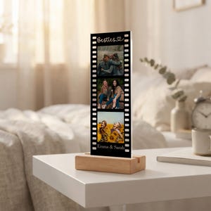 May include: A vertical rectangular acrylic photo display featuring a black film strip border design. The top text reads Besties with a heart icon, and the bottom text reads Emma & Sarah. It holds three stacked photos and sits on a light wood base.