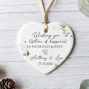 May include: A white heart-shaped ceramic ornament with a gold ribbon. The ornament has the text "Wishing you a lifetime of happiness AS HUSBAND & WIFE", names, and a date. It is decorated with floral elements, perfect for a wedding or anniversary gift.