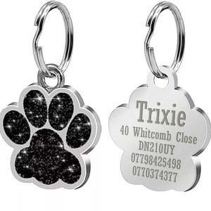 May include: Two silver dog tags. One tag is shaped like a paw print with black glitter. The other tag is a rounded rectangle with the text "Trixie", "40 Whitcomb Close", "DN210UY", "07798425498", and "0770374377".