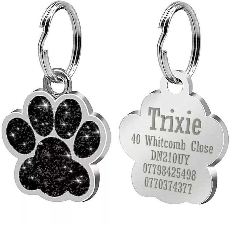 May include: Two silver dog tags. One tag is shaped like a paw print with black glitter. The other tag is a rounded rectangle with the text "Trixie", "40 Whitcomb Close", "DN210UY", "07798425498", and "0770374377".