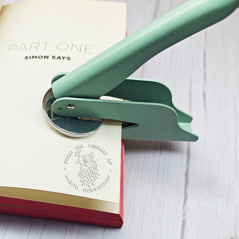 K&ouml;nnte beinhalten: Ein mintgr&uuml;ner Buchpr&auml;ger stempelt ein Buch mit dem Text "FROM THE LIBRARY OF JUDITH ROBERTSON". Das Buch ist auf einer Seite ge&ouml;ffnet, auf der "PART ONE SIMON SAYS" steht. Das Buch hat einen roten Einband mit goldenem Rand.