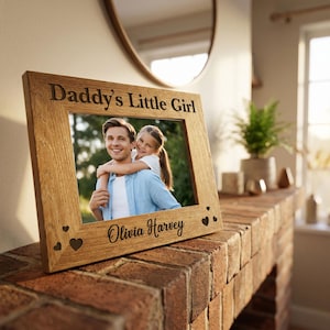 May include: A light oak picture frame with the text "Daddy's Little Girl" and "Olivia Harvey" engraved. The frame contains a photograph of a man and a girl. Heart-shaped carvings decorate the frame.