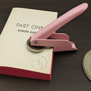 K&ouml;nnte beinhalten: Ein rosa Buchpr&auml;geger&auml;t pr&auml;gt ein Design auf eine Buchseite. Das Buch ist auf einer Seite mit "PART ONE" und "SIMON SAY" ge&ouml;ffnet. Das gepr&auml;gte Design enth&auml;lt die Initialen "S/J" in einem floralen Kranz. Das Pr&auml;geger&auml;t befindet sich auf einer dunklen Holzoberfl&auml;che.