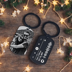 May include: Two metal keyrings with black rings. One keyring displays a black and white photo of a couple wearing sunglasses. The other keyring is black with a music waveform design and the text "em to IIA bregej nmol".