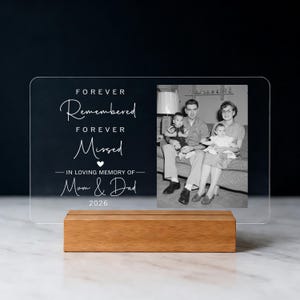 May include: A clear acrylic photo display with a wooden base. The display features a black and white family photo and the text "FOREVER Remembered FOREVER Missed - IN LOVING MEMORY OF Mum & Dad 2026".