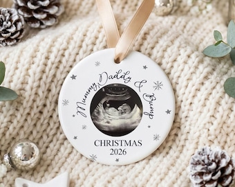 May include: A round white ceramic ornament hanging from a tan ribbon. The center features a black and white ultrasound image surrounded by the text Mummy, Daddy & Bump, with the text CHRISTMAS 2026 below and small gray stars and snowflakes.