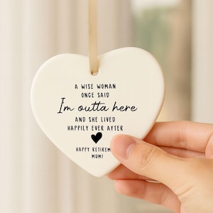 May include: A cream-coloured heart-shaped ceramic ornament. Black text reads: "A wise woman once said, I'm outta here and she lived happily ever after." A small black heart is below the text. A gold ribbon is attached for hanging. Ideal for retirement.