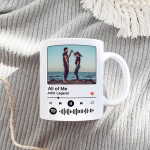 May include: White ceramic mug featuring a photograph of a couple dancing on a beach. The mug has a Spotify-style design with the song title "All of Me" by John Legend and a Spotify logo.