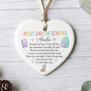 May include: A white heart-shaped ceramic ornament hanging from a gold ribbon. It features the text FIRST DAY OF SCHOOL, the name Amelia, and a poem. Illustrations include a purple notebook, a teal backpack, small hearts, and a stack of books.