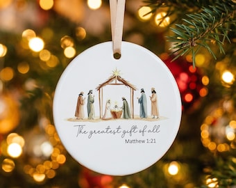 Könnte beinhalten: Ein rundes, weißes Keramikornament mit einer Aquarellillustration einer Krippenszene mit Stall, Figuren und einem Stern. Unter der Abbildung steht der Text "The greatest gift of all" und "Matthew 1:21". Ein beiges Band ist oben befestigt.