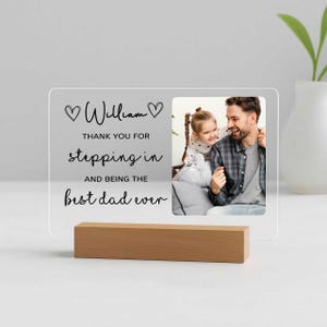 May include: A clear acrylic photo frame with a wooden base. The frame has a photo of a man and a child, and text that reads "William, Thank you for stepping in and being the best dad ever."