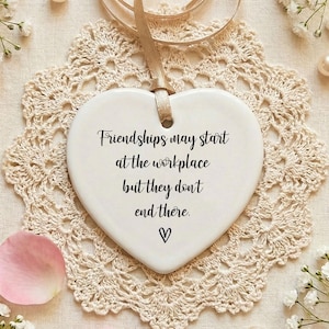 May include: A white heart-shaped ceramic ornament with the text "Friendships may start at the workplace but they don't end there." A small black heart is below the text. The ornament hangs from a beige ribbon. The ornament rests on a beige lace doily.