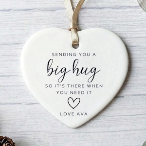 May include: White heart-shaped ornament with a gold ribbon. The ornament features the text "SENDING YOU A big hug SO IT'S THERE WHEN YOU NEED IT LOVE AVA" in black script, with a heart symbol.