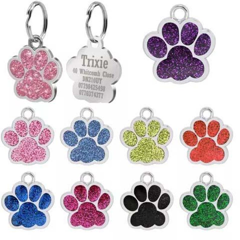May include: Ten silver dog tags with paw print designs. The tags are in various colors, including pink, blue, green, red, black, and purple. Each tag has a glitter finish. One tag has the text "Trixie 40 Whitcomb Close DN210UY 07798425498 07760374377".