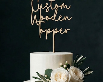 Könnte beinhalten: Ein individueller Kuchenaufsatz aus hellem Holz mit dem Schriftzug "Custom Wooden Topper" in Schreibschrift. Der Aufsatz befindet sich auf einem weißen, mehrstöckigen Kuchen, der mit weißen Rosen und Grünzeug verziert ist. Der Hintergrund ist dunkelgrün.