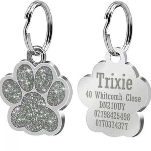 May include: Two silver dog tags with a paw print design. One tag has a sparkly paw print and the other has the text "Trixie", "40 Whitcomb Close", "DN210UY", "07798425498", and "0770374377".