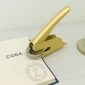K&ouml;nnte beinhalten: Ein goldfarbener Pr&auml;gestempel mit langem Griff, der auf einem Buch mit dem Wort "CODA" auf dem Einband liegt. Das Buch hat ein silbernes gepr&auml;gtes Siegel mit der Aufschrift "FROM THE LIBRARY OF ALEXANDER & BARKER".