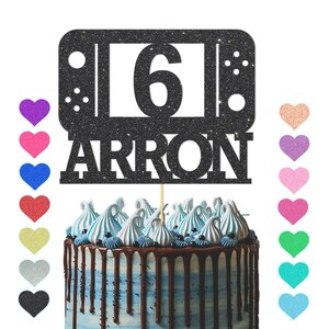 May include: Black glitter cake topper featuring a video game controller design and the number 6. The topper also includes the name "Arron".