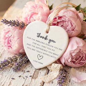 May include: A white heart-shaped ceramic plaque with the words "thank you" and a message of appreciation, along with a heart symbol and the name "Emma". The plaque is suspended by a gold ribbon and is surrounded by pink peonies and lavender sprigs.