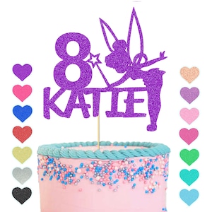 May include: Purple glitter cake topper featuring a fairy silhouette, the number 8, and the name Katie. The topper is on a pink and blue frosted cake.