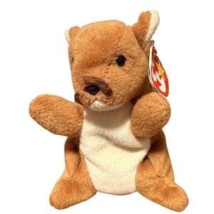 May include: A brown plush kangaroo toy with a white belly and a red and yellow tag. The kangaroo has black eyes and a brown nose. The toy is in a sitting position with its arms outstretched.
