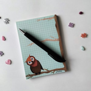 Könnte beinhalten: Ein blaues und weißes kariertes Notizbuch mit einer braunen Eulenillustration und einem schwarzen Stift. Das Notizbuch trägt den Text "©Tua Mei" am unteren Rand.