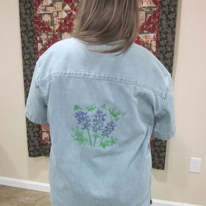 May include: A light blue denim short-sleeve button-down shirt featuring an embroidered design of purple bluebonnet flowers with green stems and leaves centered on the back panel. The shirt has a classic collar and a horizontal back yoke seam.