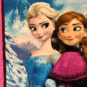 Puede incluir: Dos chicas animadas, Elsa y Anna de la pel&iacute;cula Frozen, est&aacute;n de pie una al lado de la otra. Elsa lleva un vestido azul y plateado y Anna lleva un vestido rosa y morado. El fondo es una escena de monta&ntilde;a nevada.