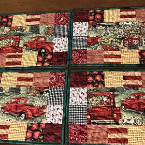 May include: Four red and green quilted placemats with a patchwork design featuring red trucks, apples, and a farmhouse.