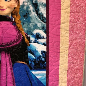 Puede incluir: Una manta acolchada rosa, crema y blanca con un estampado de personaje de Frozen. La manta presenta un primer plano de Elsa, una princesa de Disney, con un vestido rosa y azul y el pelo largo trenzado.