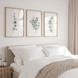 May include: A white bed with a beige knitted throw blanket and white pillows. Three framed botanical prints with green leaves and gold accents hang on the wall above the bed.