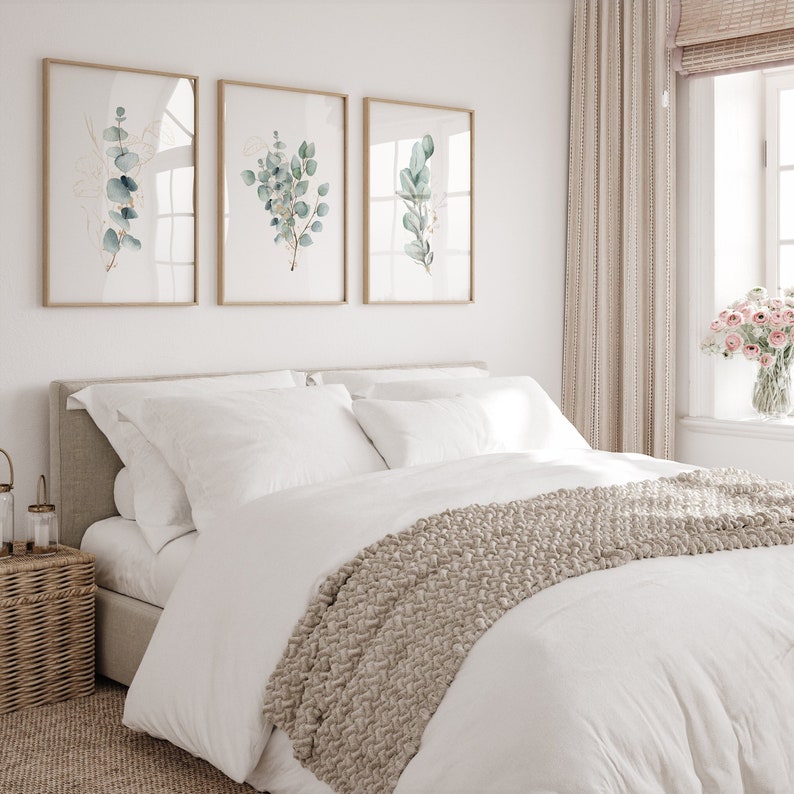 May include: A white bed with a beige knitted throw blanket and white pillows. Three framed botanical prints with green leaves and gold accents hang on the wall above the bed.