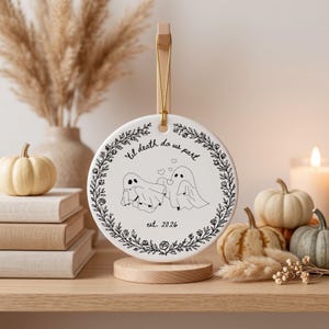 May include: A round white ceramic ornament featuring a black line drawing of two ghosts holding hands with hearts above them. The text reads 'til death do us part' and 'est. 2026' inside a circular floral wreath border. It hangs from a gold cord.