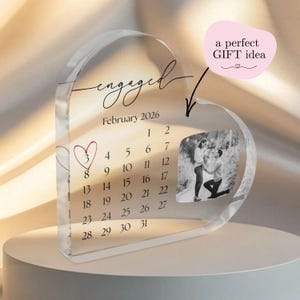 Può includere: Sottobicchiere a forma di cuore in acrilico trasparente con la scritta "engaged" e la data febbraio 2026. Presenta un calendario che evidenzia una data con un cuore e una foto in bianco e nero. È incluso anche il testo "a perfect GIFT idea".