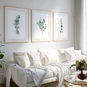 May include: Three framed botanical prints with watercolor illustrations of eucalyptus leaves in shades of green and white, hanging on a white wall above a white couch with white throw pillows and a white throw blanket.