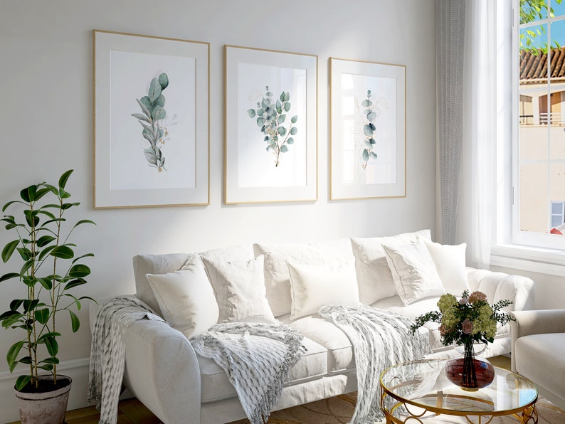 May include: Three framed botanical prints with watercolor illustrations of eucalyptus leaves in shades of green and white, hanging on a white wall above a white couch with white throw pillows and a white throw blanket.