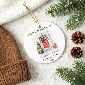 Puede incluir: Adorno navide&ntilde;o de cer&aacute;mica blanca con una cuerda dorada. El adorno presenta una ilustraci&oacute;n en acuarela de una puerta roja con una corona, un &aacute;rbol de Navidad y el texto "FIRST CHRISTMAS AT 53 HARTWAY STREET Kimberley & Jack 2025."