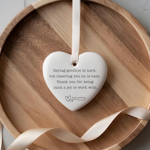 May include: A white heart-shaped ceramic ornament with a cream ribbon, inscribed with a message of appreciation. The ornament is set on a wooden tray with a cream ribbon. The text reads: "Saying goodbye is hard, but cheering you on is easy. Thank you for being such a joy to work with."