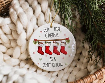 May include: A white ceramic Christmas ornament with gold string. The ornament has the text "OUR FIRST CHRISTMAS AS A FAMILY OF FOUR". Below the text are four red stockings with names. The ornament is decorated with snowflakes and greenery.