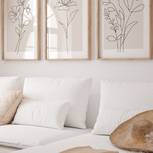 Puede incluir: Tres impresiones de arte de l&iacute;nea de flores en negro sobre un fondo beige, enmarcadas. Las impresiones est&aacute;n colgadas en una pared blanca sobre un sof&aacute; blanco con cojines beige y una manta.