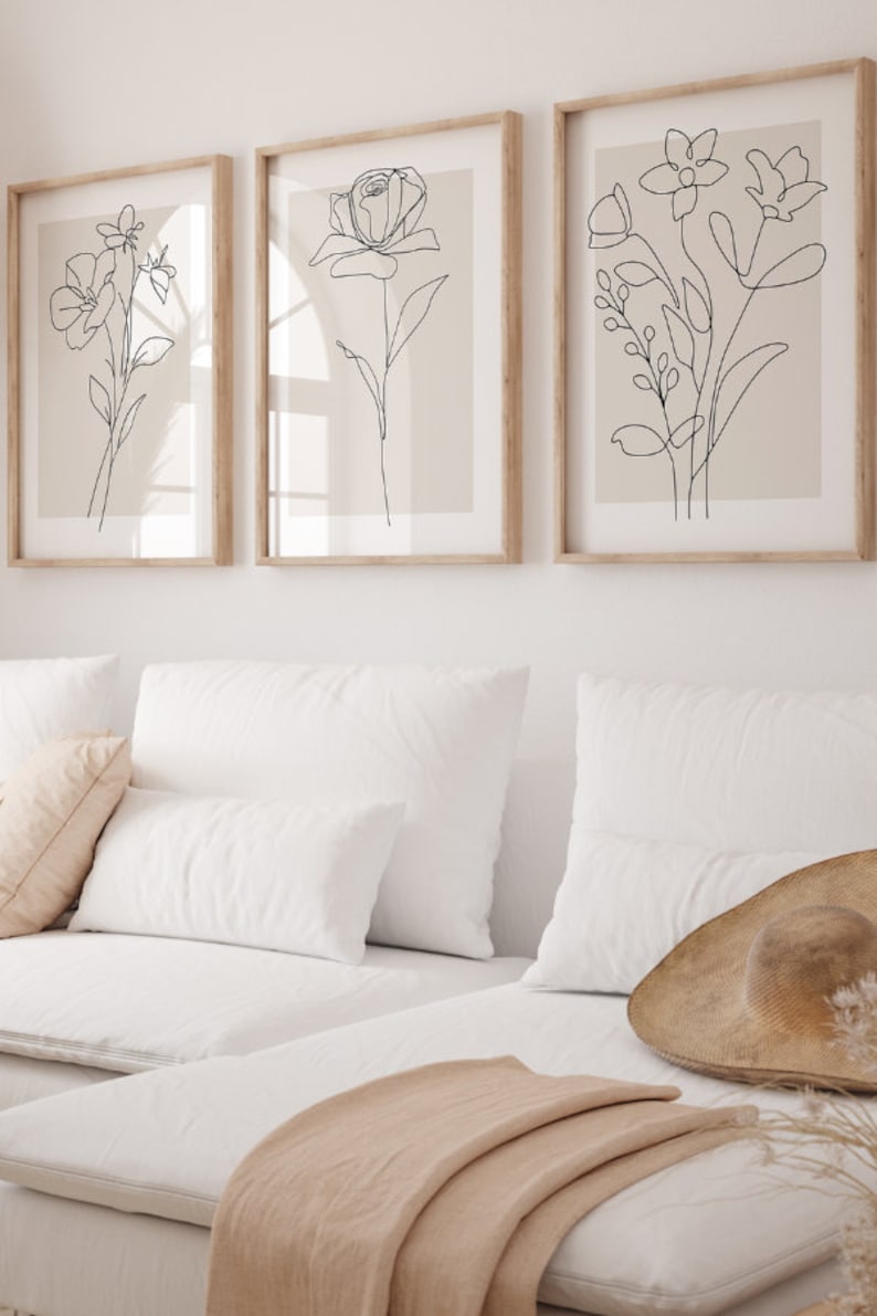 Puede incluir: Tres impresiones de arte de l&iacute;nea de flores en negro sobre un fondo beige, enmarcadas. Las impresiones est&aacute;n colgadas en una pared blanca sobre un sof&aacute; blanco con cojines beige y una manta.