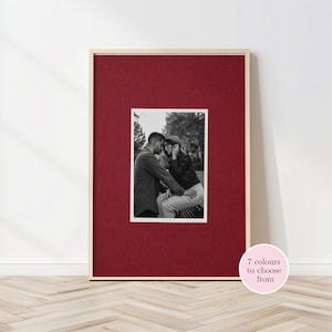 May include: A framed photograph with a black and white image of a couple. The frame is natural wood, and the mount is deep red. The text "7 colours to choose from" is in a pink circle. The frame is on a wooden floor.