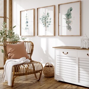 May include: A white cabinet with two drawers and two doors, a wicker chair with a brown cushion and a throw blanket, and three framed botanical prints with green leaves on a white wall.
