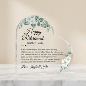 May include: A heart-shaped clear acrylic retirement gift with floral accents. The text reads "Happy Retirement Teacher Emilia" with a personalized message below. The design includes white and green floral elements.